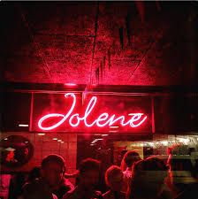 a group of people standing in front of a red neon sign that reads joliene