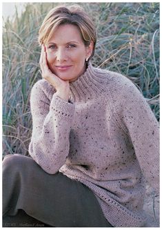 a woman sitting on the ground in front of some grass wearing a knitted sweater