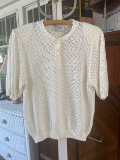 Made by D'Allaird's, this is a lovely off white/ivory acrylic sweater in an open, pointelle knit. The neckline has a decorative scalloped edging, while the short sleeves and bottom edge are ribbed. The sweater has three buttons at the neck and shoulder pads. The measurements, taken with the sweater lying flat, are: shoulder to shoulder, 17 inches; armpit to armpit, 21 inches; sleeves, 13 inches; length, 24 inches; bottom edge, 14 inches (with stretchy ribbing). The sweater overall has quite a bit of stretch. In excellent condition. Short Sleeve Knit Sweater, Fitted Vintage Short Sleeve Sweater, Vintage Short Sleeve Knit Sweater, Vintage Cream Knit Tops, Vintage Beige Cotton Sweater, Vintage Cream Sweater With Button Closure, Cream Knit Sweater, Cream Shorts, Acrylic Sweater