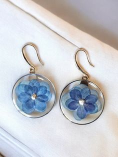 two blue flowers are hanging from earrings on a white cloth covered surface, with gold earwires in the foreground