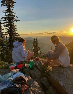 two people sitting on top of a mountain at sunset with food and drinks in their hands