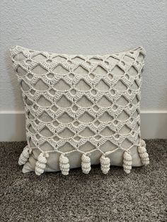 a white crocheted pillow with tassels on the bottom and sides, sitting against a gray wall