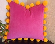 a pink pillow with yellow pom - poms sitting on a table next to a potted plant