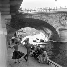 black and white photograph of people sitting on the side of a river under a bridge