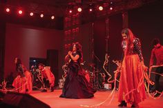 two women in red dresses are on stage with other performers behind them and one woman is holding a microphone