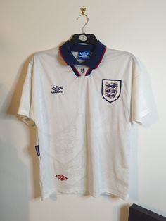 This classic England home shirt from 1993-95 is a must-have for any football fan. With the iconic Umbro branding and short sleeves, it's the perfect way to show your support for the national team. Made for adults in size medium, this shirt is suitable for both casual wear and for display as a sports memorabilia item. The white design with the red and navy accents make it a timeless piece that will never go out of style. Whether you are a diehard supporter of the Three Lions or just appreciate th White Shirt With Team Logo For Team Spirit, White Team Spirit Shirt With Logo, White Shirt With Team Logo, White Collegiate Shirt For Fan Merchandise, White Shirt With Team Logo For Sports Season, White Team Logo Shirt For Sports Events, White Shirt With Team Logo For Sports Events, White Team Logo Shirt For Fan Merchandise, White Shirt With Team Logo For Fans