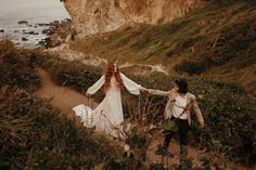 a man and woman walking down a dirt path next to the ocean with their arms around each other