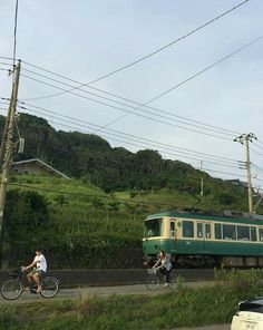 two bicyclists ride past a green train on the tracks near a grassy hill