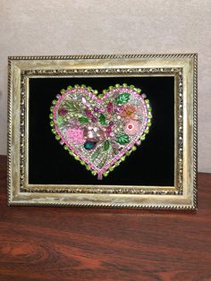 a heart made out of beads in a gold frame on a wooden table next to a wall