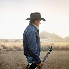 a man wearing a cowboy hat and holding a guitar in the middle of a desert