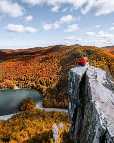 a man sitting on top of a cliff next to a lake in the middle of autumn