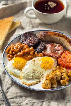 an english breakfast with sausage, eggs, beans and tomatoes on a plate next to a cup of tea