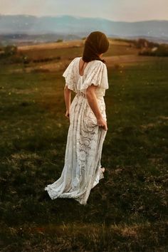 a woman in a white dress standing on top of a lush green field with mountains in the background