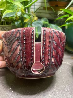 a hand holding a red and black bowl on top of a table next to potted plants