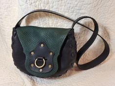 "This leather bag is made from a 1970's pattern that we dug up, it has a cool retro feel but also works well for steampunk or modern style! It is made of black and green bullhide with black cowhide for the bottom gusset, front detail, and strap.  It is completely hand stitched with our own very strong leather cording that we make, so this is a very sturdy bag.  This one has a decorative piece on the front flap with antiqued rivets and a slot which has a brass ring that passes through to hold it closed.  The bag is 9\" wide at the widest point, 8\" tall, and the bottom is about 3.5\" deep. The strap is 2\" wide and 38\" long, can be worn on the shoulder or across the body." Handmade Leather Shoulder Bag, Brass Ring, Rivets, Leather Cord, Leather Shoulder Bag, Leather Handmade, Hand Stitching, Purses And Handbags, Leather Bag