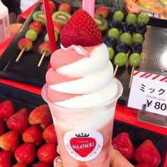 a person holding up a cup with ice cream and strawberries on it in front of some fruit