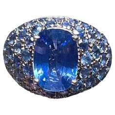 This is an 18K white gold blue sapphires dome cocktail ring. It has an oval cut faceted blue sapphire prong setting in the center, whose measurements are 11.98 x 8.35 x 4.58mm and its weight is approximately 3.78 carat. Pave cluster of fifty one diamond cut sapphires that each one measures 3.0 mm and weights 0.12 carats enhance this queenly ring. It is stamped 750 inside the ring. Domed Ring, Gem Stone, Cocktail Ring, Cocktail Rings, Oval Cut, Prong Setting, Blue Sapphire, Diamond Cuts, Jewelry Rings