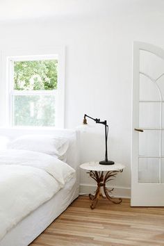 a bedroom with white walls and wood floors, an arched window over the bed is next to a small table with a lamp on it