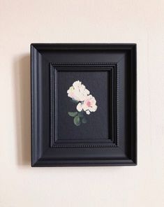 a black frame with white and pink flowers hanging on the wall next to a shelf