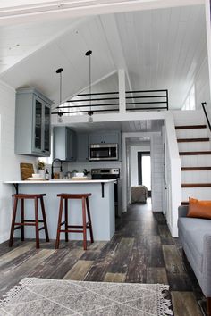 a living room and kitchen area in a tiny home with wood floors, white walls, gray cabinets, and wooden bar stools