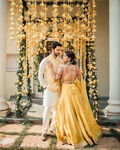 a man and woman standing in front of a yellow flower covered archway with flowers hanging from the ceiling