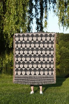 a woman standing in the grass holding up a black and white quilt with trees on it