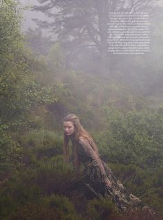 a woman with long hair is sitting in the grass and trees on a foggy day