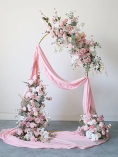 a pink and white wedding arch with flowers on the top, draped over by a pink ribbon