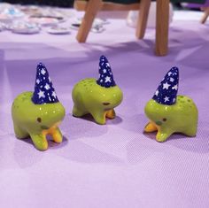 three green elephants with blue hats on their heads sitting on a purple cloth covered table