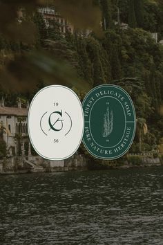 two logos on the side of a body of water with houses in the background and trees behind them