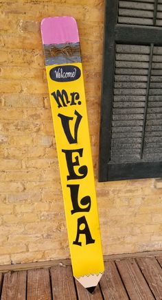 a yellow and pink sign that says welcome to me in black writing on it, next to a brick wall