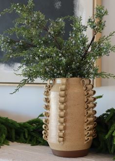 a vase filled with lots of green plants on top of a table next to a painting