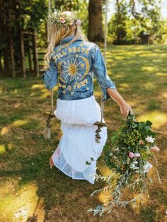 a woman in a white dress and jean jacket is walking through the grass with flowers