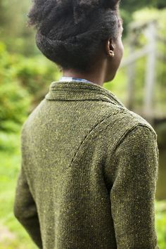 a woman standing in the woods with her back to the camera, wearing a green sweater