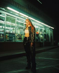 a woman standing on the sidewalk in front of a store at night with neon lights