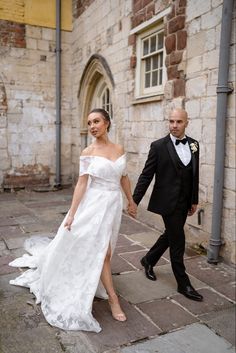 a bride and groom walking down the street holding hands in front of an old brick building