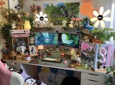 a desk with two computer monitors on top of it next to plants and stuffed animals