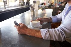 a man sitting at a table using his cell phone while holding a cup of coffee