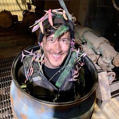 a man wearing a hat with ribbons around his head sitting in a metal container filled with junk