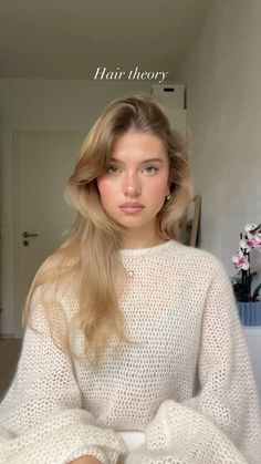 a woman with long blonde hair sitting in front of a mirror wearing a white sweater