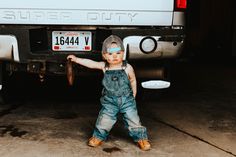 a little boy in overalls and a hat standing next to a white pickup truck