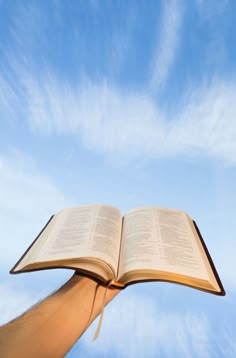 an open book being held up in the air by someone's hand, against a blue sky with wispy clouds