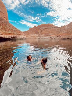 two people are swimming in the water near some mountains and cliffs, with blue skies above them