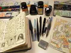 many different types of pens and ink on a table next to an open book with drawings