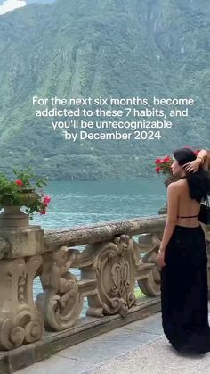 a woman in a long black dress standing on a balcony next to flowers and mountains