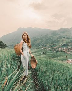 a woman standing in tall grass with a basket on her shoulder and mountains in the background