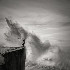 a man standing on top of a cliff while holding onto a surfboard in front of a huge wave