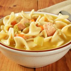 a white bowl filled with pasta and meat soup on top of a wooden table next to a spoon