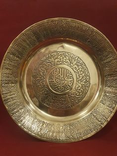 an ornate gold plate with arabic writing on the front and sides, sitting on a red surface