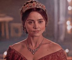 a woman in a red dress wearing a gold tiara and diamond necklace with candles behind her